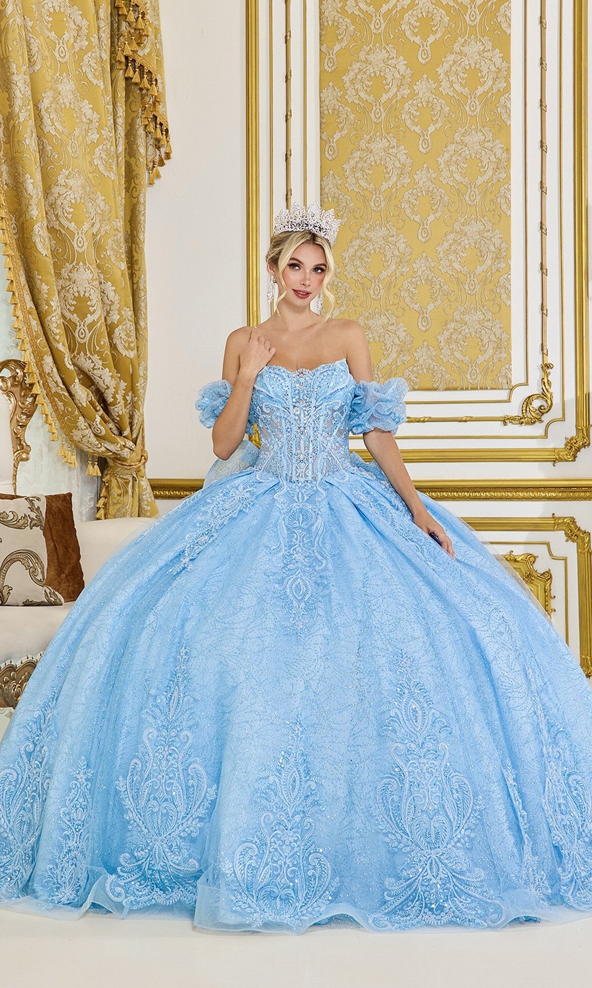 A woman wearing the Quinceanera Dress 5100 by Dancing Queen in dusty blue poses with a jeweled crown in an elegant, gold-accented room featuring patterned wallpaper and drapes.